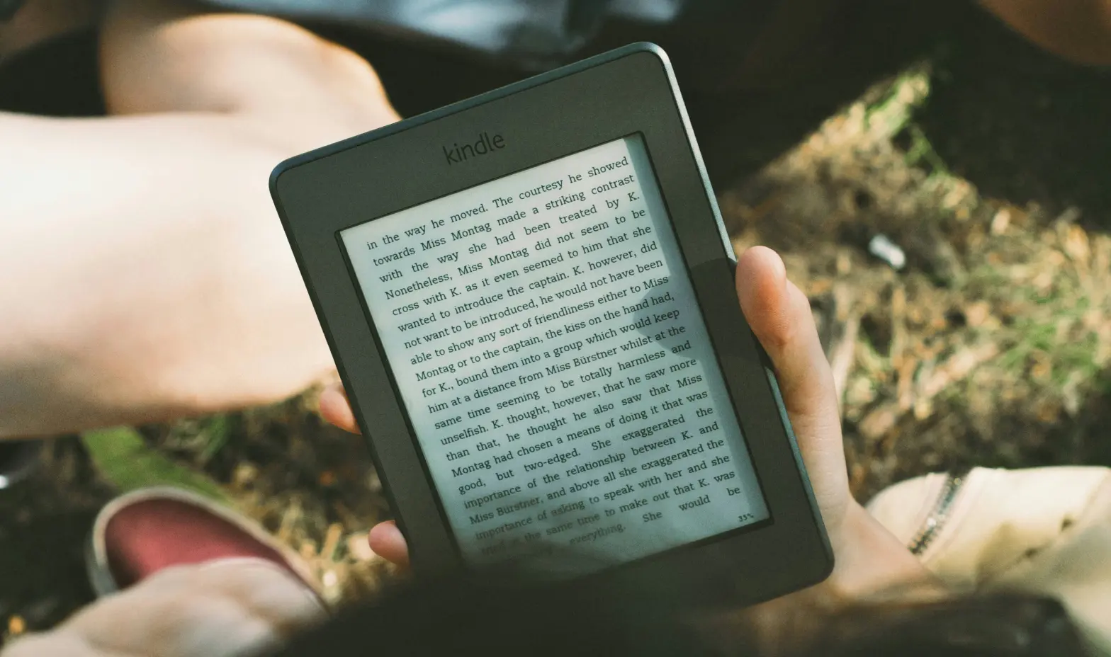 A person relaxing in the grass, holding a Kindle and enjoying a good book under the sun.