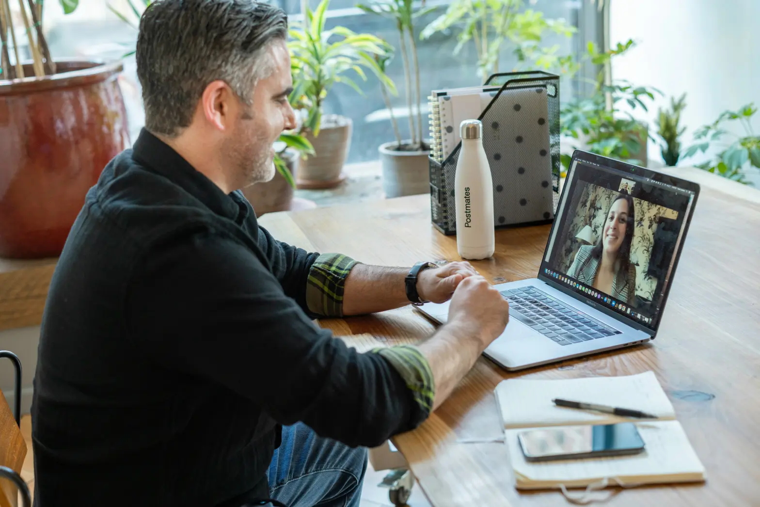 Man sitting at a desk using a laptop for a video call with a woman on screen. Desk has a notebook, pen, phone, and a water bottle. Plants are in the background.