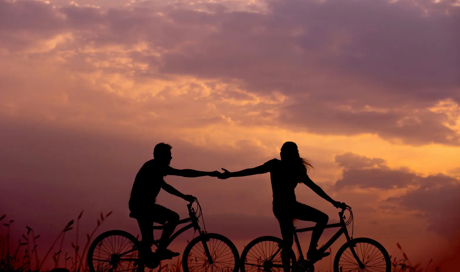 Silhouettes of two people on bicycles holding hands at sunset under a cloudy sky.
