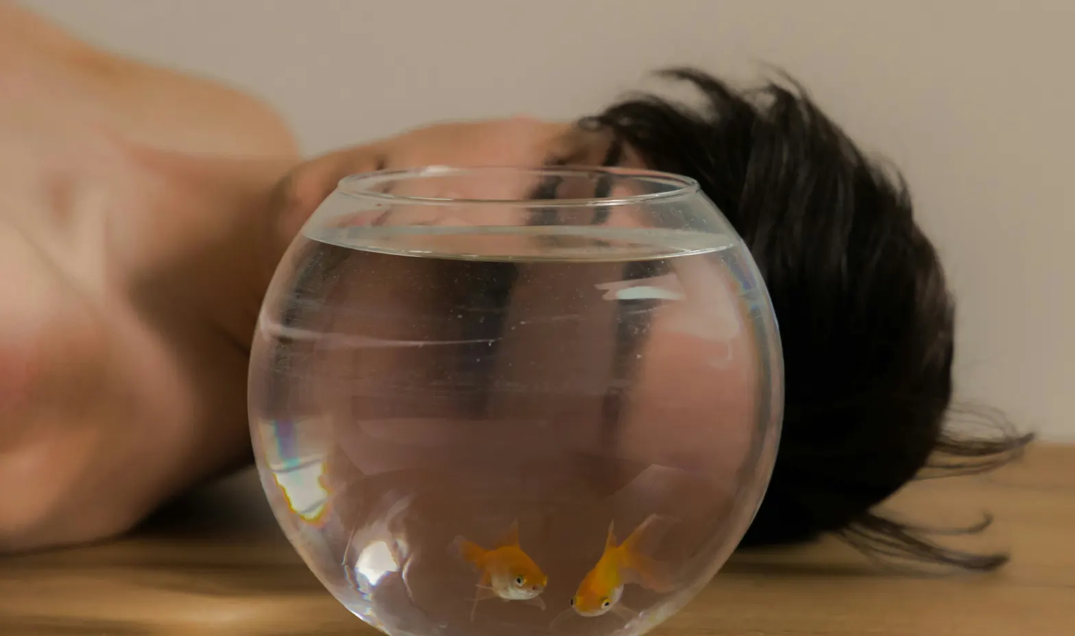 A man reclines on a table beside a fish bowl, creating a quirky and relaxed scene.