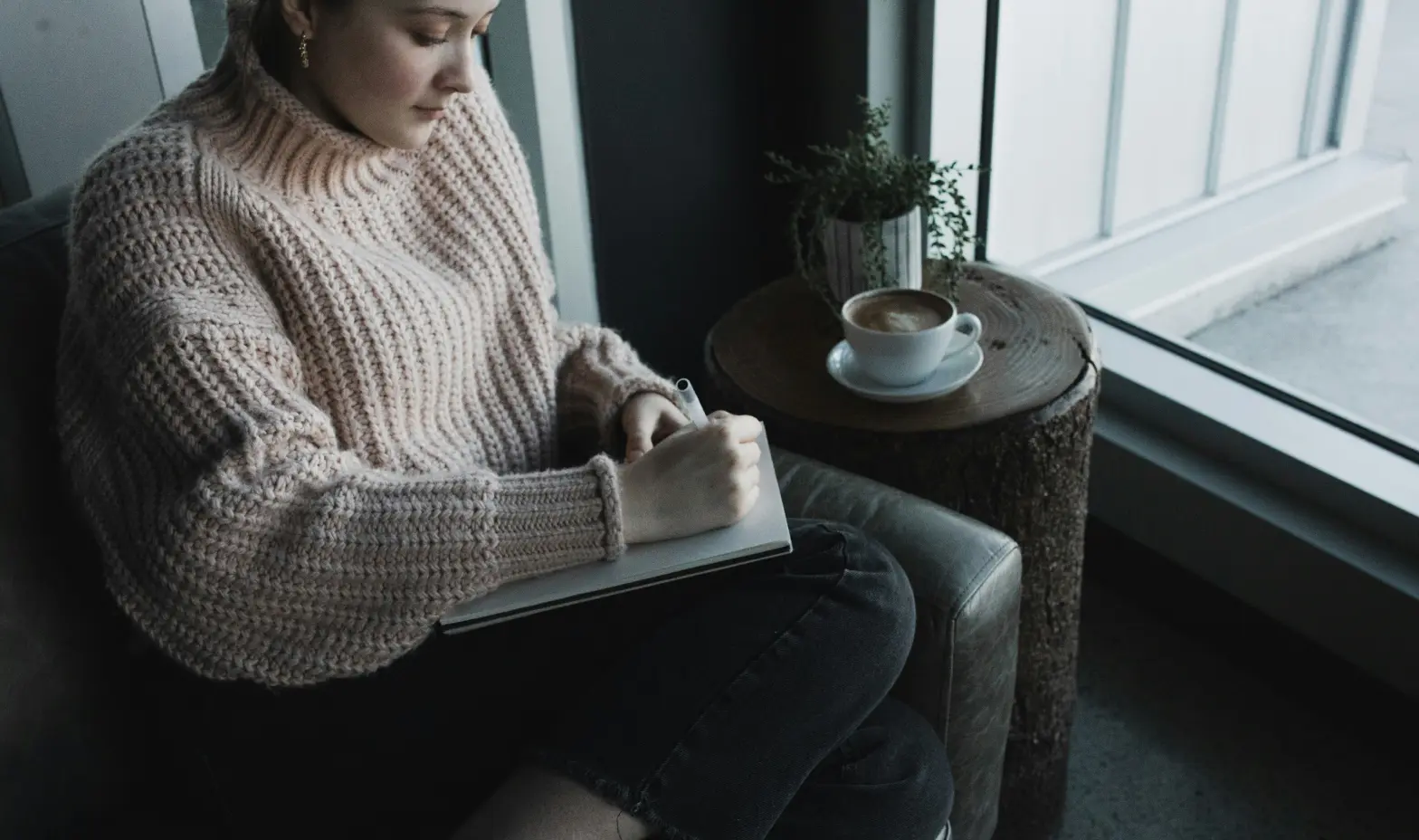 A woman in a cozy sweater sits on a couch, focused on writing in her notebook.