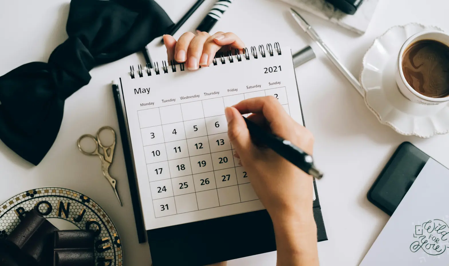 A woman's hand notes on a calendar, accompanied by a pen and a warm cup of coffee.