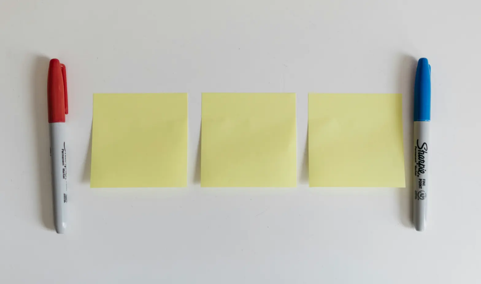 A trio of sticky notes in vibrant colors on a white background, with two pens placed nearby for writing.