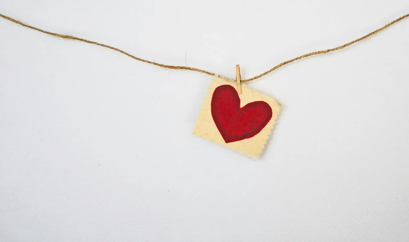 A small red heart on a piece of beige paper hangs from a string with a clothespin against a plain white background.
