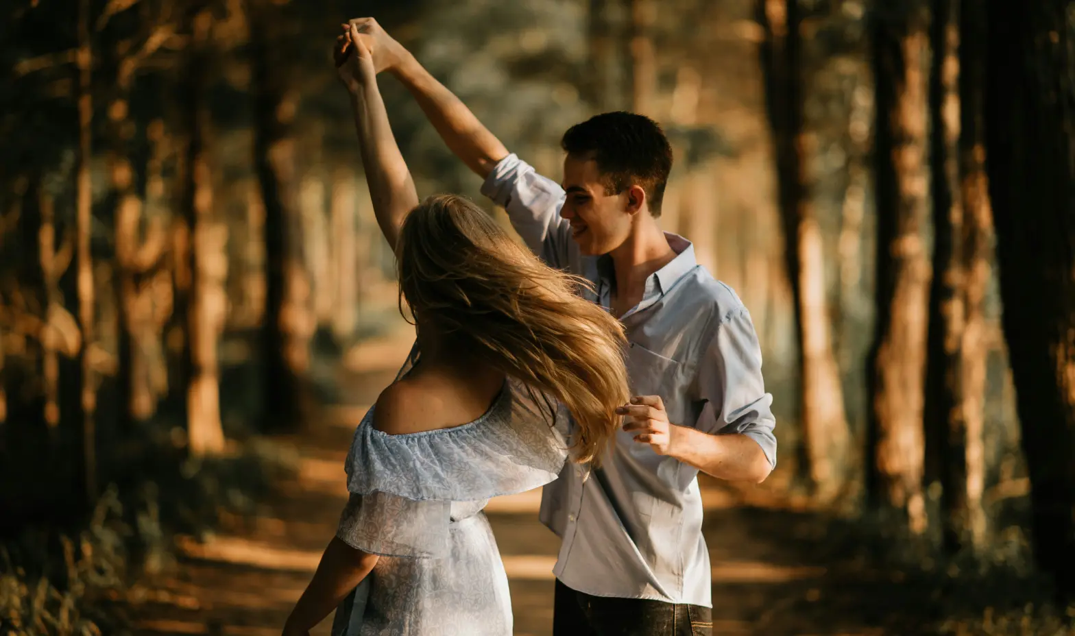 A happy young couple twirls in the woods, surrounded by greenery and soft sunlight filtering through the trees.