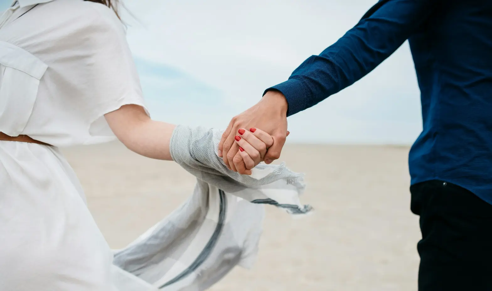 A man and woman holding hands, walking together on the beach sand, enjoying a sunny day.