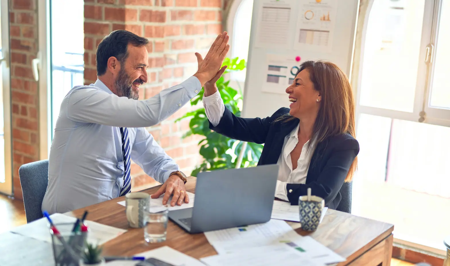A man and woman in business attire share a joyful high-five in a bright office.