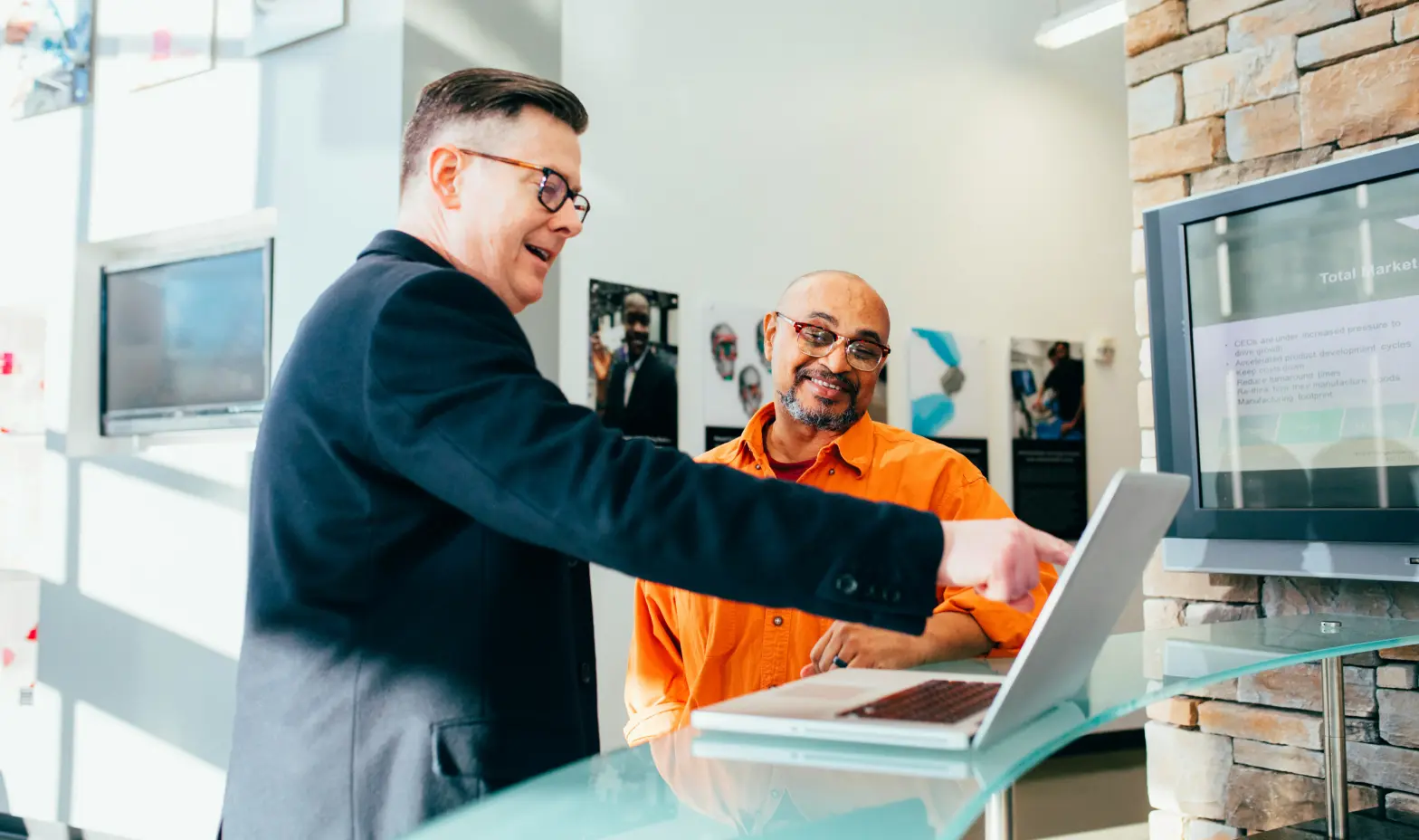 Two men standing at a reception desk with a laptop. One is pointing at the screen, while the other smiles.