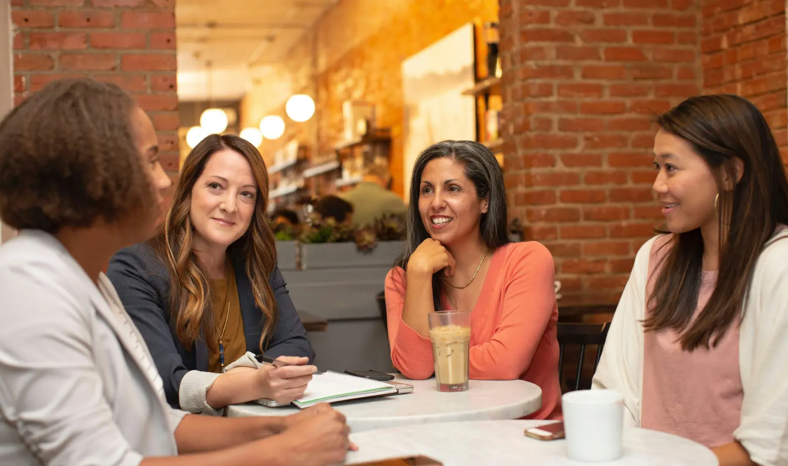 Four women sit around a table in a cozy cafe with brick walls, engaged in a friendly conversation.