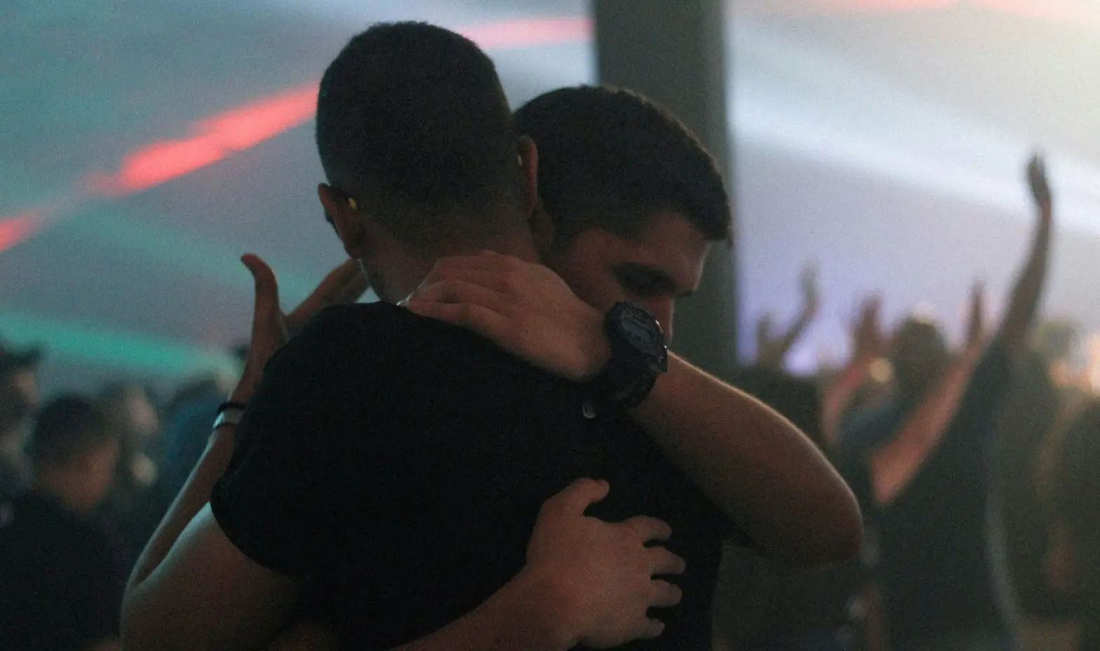 Two men share a warm hug in a lively nightclub, surrounded by colorful lights and a vibrant atmosphere.
