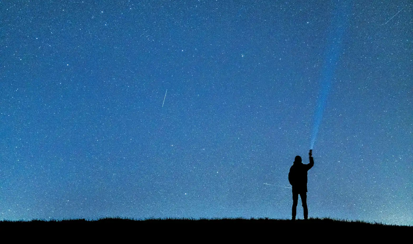Silhouette of a person on a hill at night holding a lantern pointed toward a starry sky with meteors.