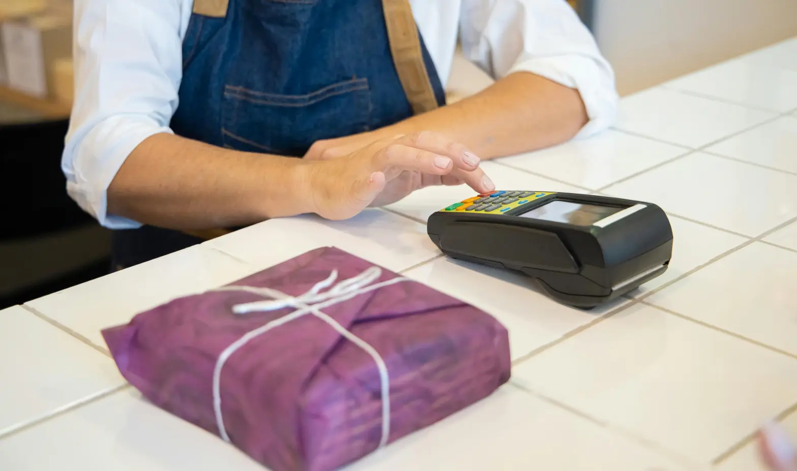 A person in an apron stands at a counter, using a credit card machine to process a payment.