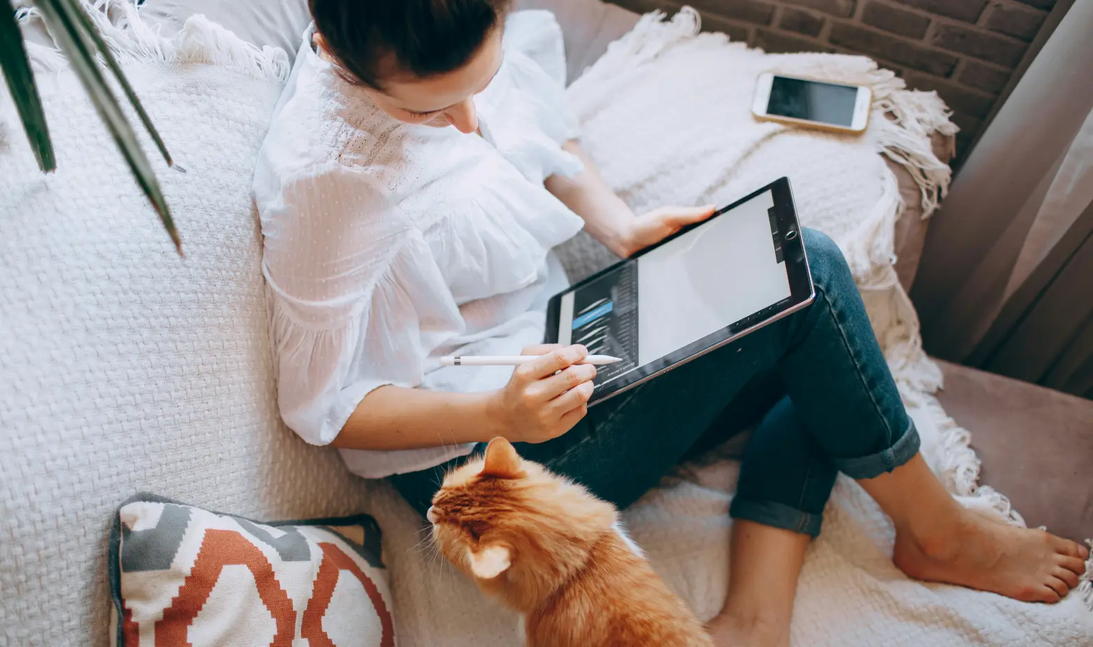 A woman sits on a couch with a cat in her lap, focused on her laptop.