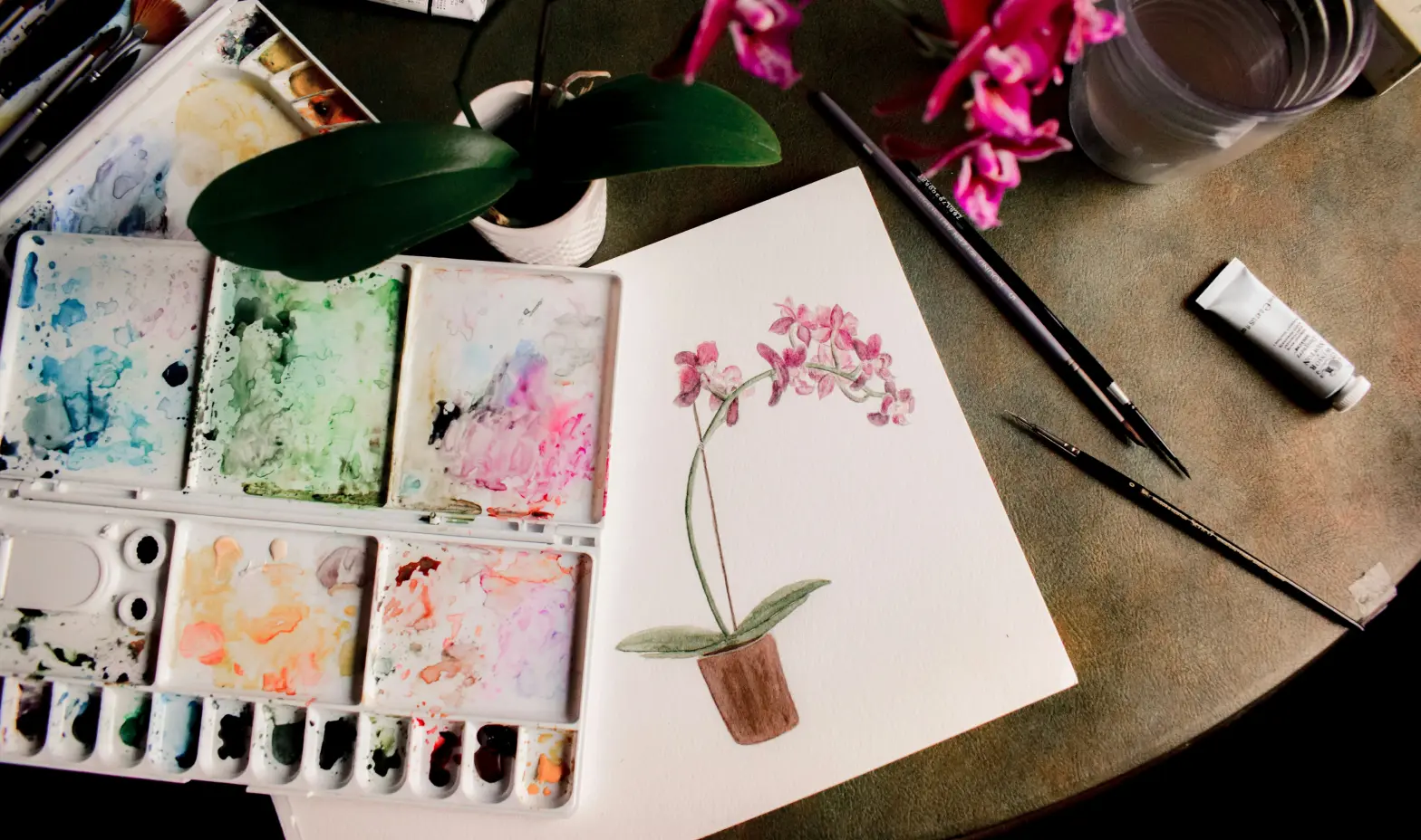 Watercolor painting of pink orchids in a pot, artist's supplies nearby.