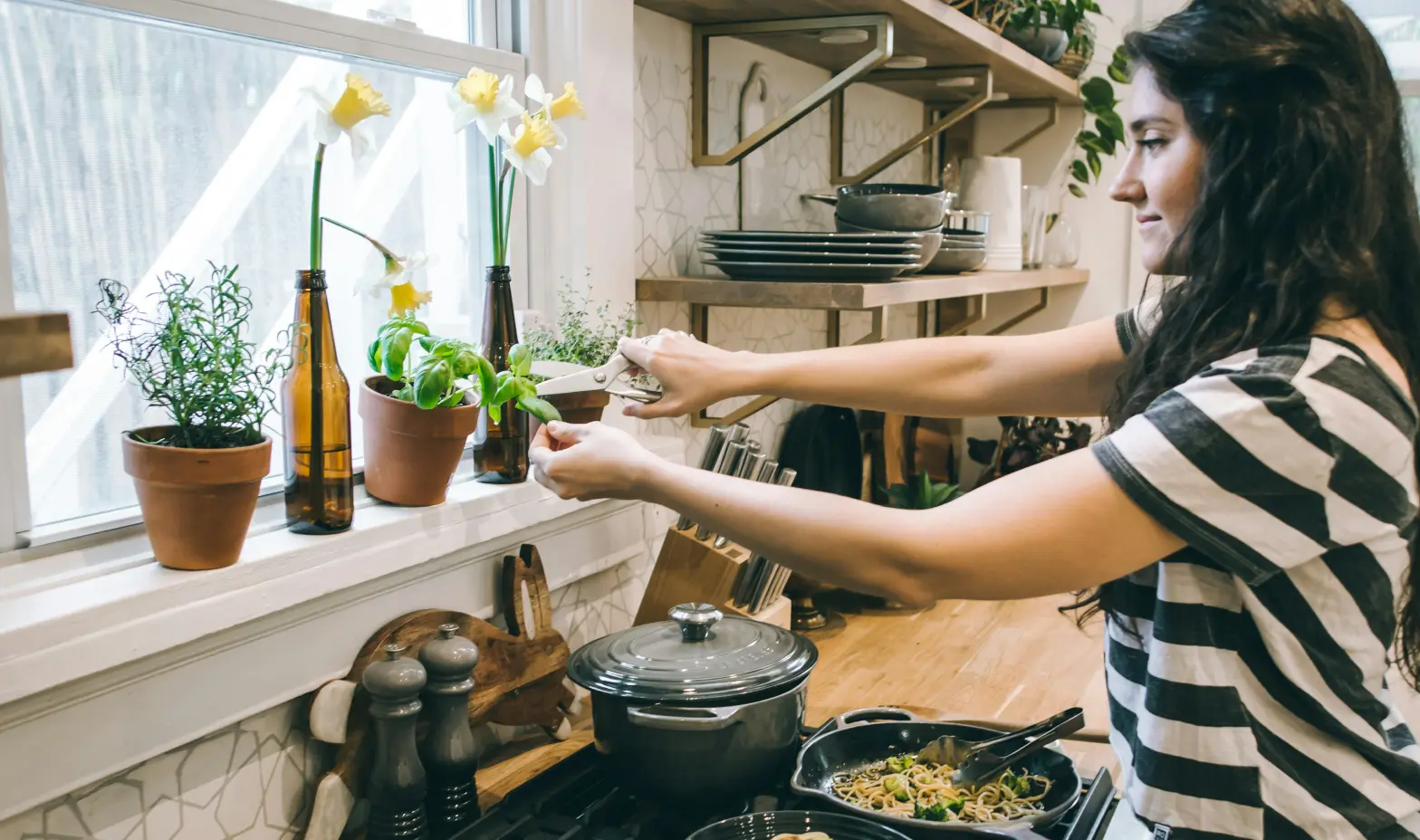 Woman cooking pasta with broccoli in a kitchen, herbs and flowers on windowsill.