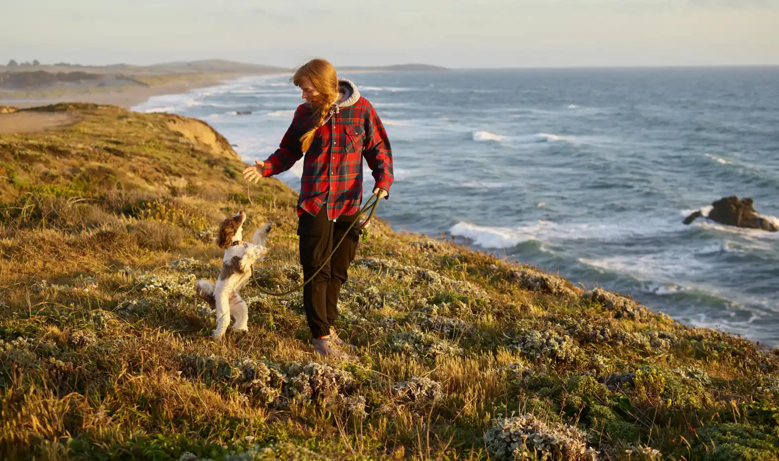 Woman playing with her dog on a coastal cliff at sunset.