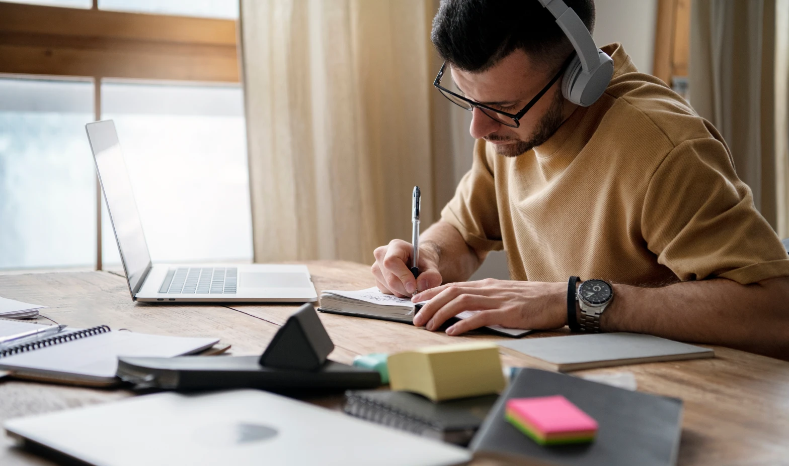 A man in headphones works intently on his laptop, taking notes in a notebook.
